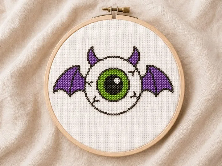 Flying eyeball with purple bat wings cross stitch pattern stitched and shown in an embroidery hoop